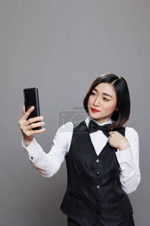 Photo for Smiling woman receptionist chatting in remote conversation on mobile phone. Young attractive asian waitress looking at smartphone front camera while taking selfie and posing in studio - Royalty Free Image