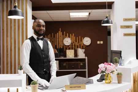 Photo for African american bellboy works at hotel, ensuring professional luxury concierge services for guests. Male resort employee providing assistance at check in, helping with accommodation. - Royalty Free Image
