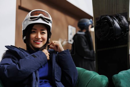 Photo for Female asian traveler sits on hotel lobby couch, wearing winter jacket and adjusting her ski goggles. Woman modifies her snow helmet for safety when snowboarding at slopes of the luxury ski resort. - Royalty Free Image