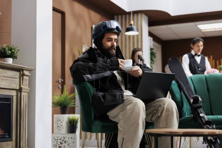 Photo for In luxury lounge area, man dressed in winter jacket enjoys coffee while using laptop to explore winter activities. Combination of technology and comfort adds excitement for tourist with skiing gear. - Royalty Free Image