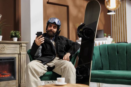Téléchargez les photos : Homme avec des vêtements d'hiver parlant sur smartphone à la réception de l'hôtel assis avec du matériel de snowboard près de la cheminée. Voyageur en équipement de neige en utilisant un appareil mobile dans le salon de la station de ski. - en image libre de droit