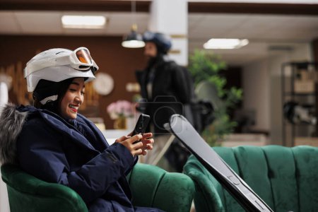 Photo for Side-view of woman using mobile device in hotel lobby, engrossed in surfing the net. Tourist holding cell phone, equipped with winter jacket, ski goggles and skiing skis for fun winter holiday. - Royalty Free Image
