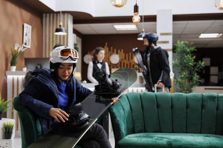 Photo for Young female guest carrying and checking her snowboard in lounge area on winter mountain resort. Asian woman seated in hotel reception and maintaining her snowboarding equipment. - Royalty Free Image