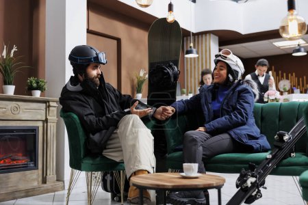 Téléchargez les photos : Couple vêtu de vêtements d'hiver chauds assis et parlant sur un canapé confortable dans le hall de l'hôtel. Des hommes et des femmes excités portant des lunettes de ski et des vestes d'hiver se tiennent la main, du matériel de ski appuyé sur le canapé. - en image libre de droit