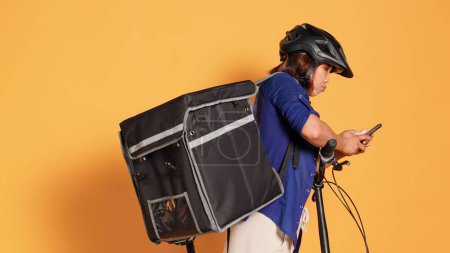 Photo for Handheld camera shot of courier texting client after delivering wrong takeout order. Bike rider wearing safety helmet and thermic bag holding food, isolated over studio background - Royalty Free Image