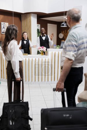 Téléchargez les photos : Couple âgé caucasien entre dans l'hôtel et s'approche de la réception pour l'assistance de réservation des employés multiethniques. Les réceptionnistes professionnels saluent les retraités avec des sourires. - en image libre de droit