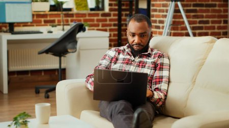 Photo for Easy going african american employee working from home on laptop, Teleworker remotely doing various tasks, typing on keyboard in stylish cozy warm apartment personal office - Royalty Free Image