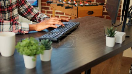 Photo for Employee remotely working from home on computer, inputing data in his brick wall apartment. African american freelancer typing on keyboard, teleworking at nice clean wooden desk, close up - Royalty Free Image
