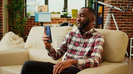 Foto de Hombre afroamericano disfrutando de un día libre en casa, charlando con amigos en videollamada. Sonriente persona alegre emocionado de ponerse al día con los compañeros durante la reunión en línea mientras se relaja en acogedor apartamento acogedor - Imagen libre de derechos