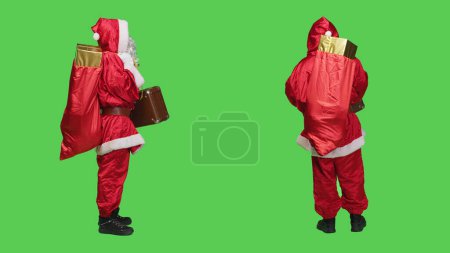 Foto de Saint Nick con bolsa de regalos y maletín, en busca de transporte para entregar regalos a los niños de todo el mundo. Joven retratando a Santa Claus con maleta vintage y saco rojo. - Imagen libre de derechos