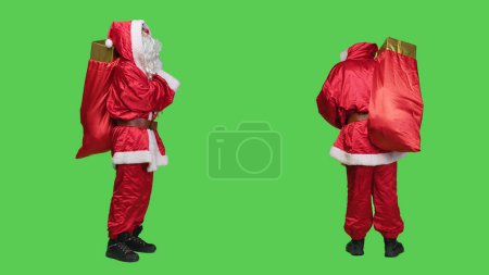 Photo for Saink nick in suit looking around, carrying big red bag full of gift boxes over full body greenscreen backdrop. Young person dressed as santa spreading christmas holiday spirit, presents for kids. - Royalty Free Image