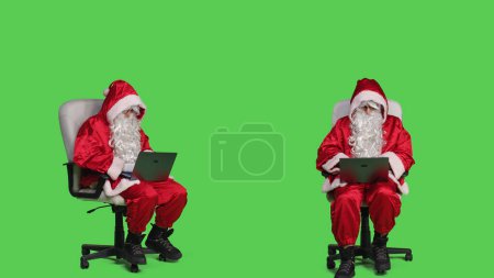 Foto de Modern saint nick trabaja en el ordenador portátil en el estudio, sentado en la silla contra el fondo de pantalla verde de cuerpo completo. Carácter de Santa Claus con PC portátil inalámbrica, navegar por el sitio en línea en traje festivo. - Imagen libre de derechos