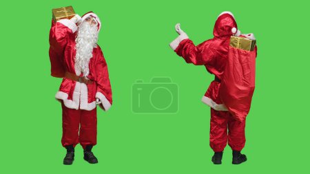 Téléchargez les photos : Santa Claude avec sac rouge dit ho ho ho sur fond plein corps vert, portant costume de vacances traditionnel et des lunettes. Père Noël cosplay portant sac avec des cadeaux et des jouets. - en image libre de droit