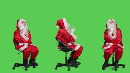 Foto de Papá Noel pensativo en traje se sienta en la silla, pensando en las ideas de regalo de Nochebuena sobre pantalla verde de cuerpo completo. Joven actuando como santo nick carácter con barba blanca, lluvia de ideas. - Imagen libre de derechos