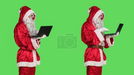 Téléchargez les photos : Père Noël homme sur videocall en ligne agitant à l'écran de l'ordinateur portable pour discuter avec les gens sur les cadeaux, debout sur fond d'écran vert. Personne en costume de fête rouge par téléconférence. - en image libre de droit