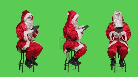 Photo for Santa working on digital tablet during christmas eve holiday, expressing december seasonal event. Young man saint nick sitting on chair against full body greenscreen backdrop, using gadget. - Royalty Free Image