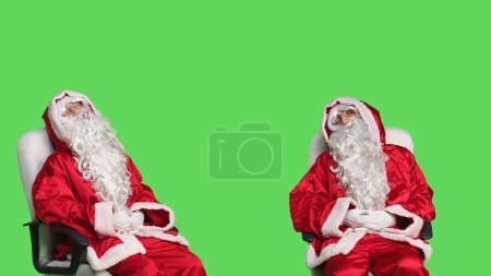 Téléchargez les photos : Père Noël s'endort sur une chaise, essayant de garder les yeux ouverts en studio avec écran vert. Homme en costume saint pseudo dormir à la caméra, se sentir fatigué et relaxant avant la célébration festive. - en image libre de droit