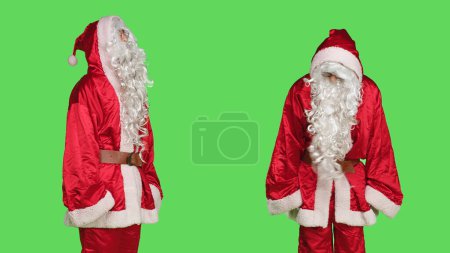 Photo for Young man in santa claus suit saying no posing over greenscreen backdrop, acting displeased while he wears traditional winter costume. Negative person in saint nick cosplay, disappointed santa. - Royalty Free Image