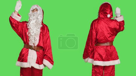 Photo for Saint nick enbodiment greeting people, wearing festive seasonal costume and waving hello on greenscreen backdrop. Young adult celebrating christmas eve in red suit, smiling at camera. - Royalty Free Image