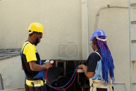 Photo for African american coworkers hired after complaints about noisy air conditioner and significant increase in energy bills. Engineers using professional manifold gauges to detect optimum fixing method - Royalty Free Image