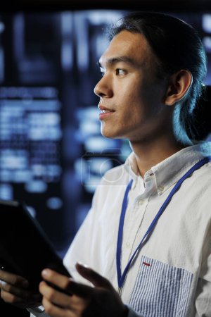 Photo for Developer in high tech facility uses tablet to prevent system overload during peak traffic periods. Asian programmer between server rows ensuring enough network bandwidth for smooth operations - Royalty Free Image