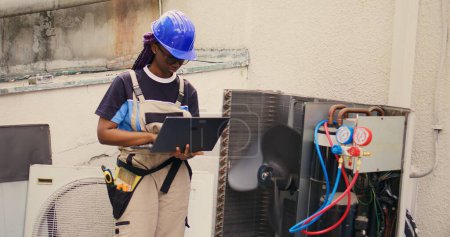 Téléchargez les photos : Électricien afro-américain qualifié travaillant sur un système hvac extérieur, tenant un ordinateur portable. Serviteur agréé optimisant les performances du climatiseur externe, s'assurant qu'il fonctionne à sa capacité maximale - en image libre de droit
