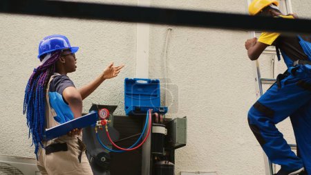 Téléchargez les photos : Precise worker measuring refrigerant levels in air conditioner with professional manometers while coworker steps down from folding ladder after finishing checking rooftop HVAC system - en image libre de droit