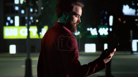 Photo for Young man on nighttime walk using smartphone, looking around city center to admire downtown district tall buildings. Business person on phone app strolling under streetlights. Handheld shot. - Royalty Free Image