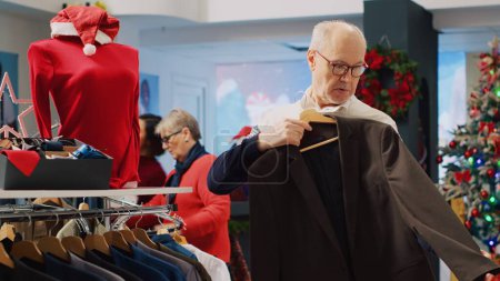 Photo for Senior man browsing blazers in festive clothing store during winter holiday season, looking at formal attire pieces, doing Christmas shopping spree in xmas decorated shop - Royalty Free Image