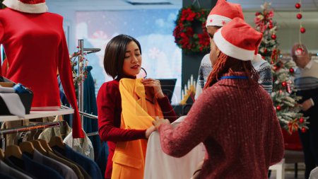 Téléchargez les photos : Assistant de vente au détail asiatique et collègue aidant client indécis choisir entre deux couleurs de blazers dans la boutique de vêtements décorés de Noël. Employés aidant le client pendant les vacances d'hiver - en image libre de droit