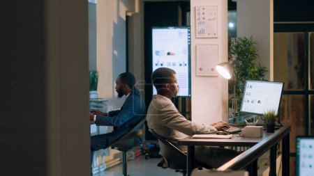 Téléchargez les photos : Superviseur afro-américain demandant au comptable de lui montrer les progrès des tâches du projet d'entreprise dans le bureau moderne pendant le quart de nuit. Cadres supérieurs supervisant les employés travaillant sur ordinateur - en image libre de droit