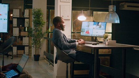 Téléchargez les photos : Un employé afro-américain en poste tient une téléconférence d'équipe avec des collègues travaillant à domicile. Homme d'affaires bavarder avec le personnel de l'entreprise pendant le quart de nuit vidéocall en ligne - en image libre de droit