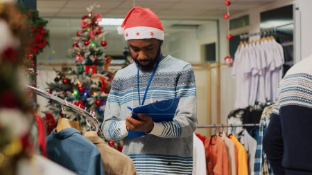Foto de Empleado afroamericano navegando a través de estantes de ropa en la tienda de moda decorada de Navidad, anotando los precios ajustados en el portapapeles. Trabajador inspeccionando blazers, buscando daños - Imagen libre de derechos