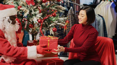 Téléchargez les photos : Client dans un magasin de mode parlant avec l'acteur habillé en Père Noël, assis à côté de l'arbre de Noël lors d'un événement promotionnel festif. Client recevant un cadeau du travailleur pendant la période des Fêtes - en image libre de droit