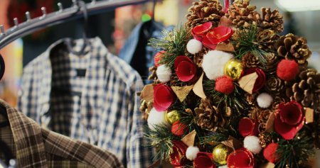 Téléchargez les photos : Dolly out shot of festive Christmas wreath decorated with pine cones hanging from clothes rack in empty shopping mall store, ready to bring holiday cheer during winter holiday season, close up - en image libre de droit