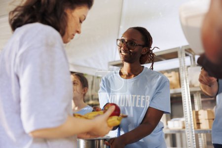 Photo for Portrait of female charity worker handing out fresh fruits and food to poor and homeless people. Cheerful black woman volunteers at food drive serving free meals to the hungry and less fortunate. - Royalty Free Image