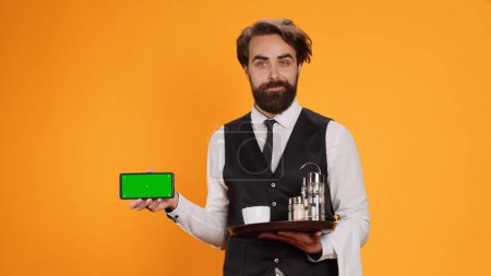 Photo for Staff member points at greenscreen on phone, showing isolated mockup template on display against yellow background. Experienced waiter presenting empty copyspace modern telephone on camera. - Royalty Free Image