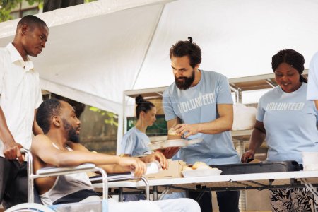 Freundliche kaukasische Hilfskräfte servieren dem Rollstuhlfahrer eine warme Mahlzeit in der Outdoor-Food-Bank. Junge Freiwillige helfen armen und weniger begünstigten afrikanisch-amerikanischen Männern und spenden kostenlose Lebensmittel.