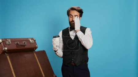 Photo for Doorman holding coffee drink in studio, wearing white gloves and feeling confident working in hotel industry. Bellhop worker in suit and tie drinking beverage next to trolley bags. - Royalty Free Image