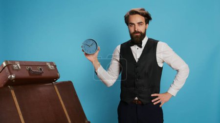 Photo for Hotel employee checking time on watch, working as bellboy or doorkeeper and holding wall clock to look at hour. Valet in formal clothing posing next to trolley bags, trying to be punctual. - Royalty Free Image