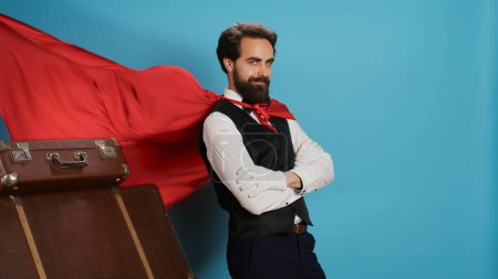 Téléchargez les photos : Travailleur de l'hôtel avec manteau rouge pour aider les clients avec des sacs trolley, prétendant être personnage de super-héros de dessin animé fantaisie à la caméra. Bellhop agissant comme un surhumain comique avec cape sur fond. - en image libre de droit