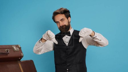 Photo for Staff member acts unhappy giving thumbs down in studio, dressed in suit. Experienced bellboy showcasing negative emotions of rejection and disappointment that lead to sadness, displeased bellhop. - Royalty Free Image