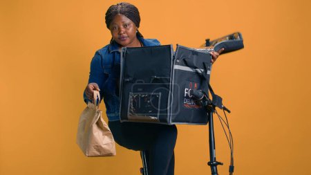 Photo for African american woman grabbing takeaway from food delivery bag for person in local neighborhood. Smiling courier providing fast and reliable customer service by giving fresh takeout meal to client. - Royalty Free Image