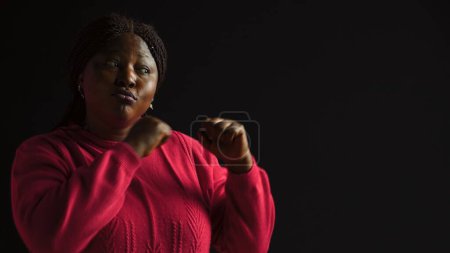 Photo for African american woman radiating confidence stands with hands clasped ready for challenge. Young lady poses with clenched fists embodying strong and self-assured presence in isolated black background. - Royalty Free Image