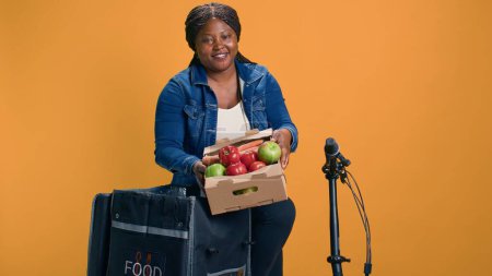 La mensajera afroamericana transporta suavemente la cesta de fruta de la bolsa de entrega de alimentos. Mujer partera emocionada en su bicicleta agarrando una caja de productos frescos y saludables para entregar.