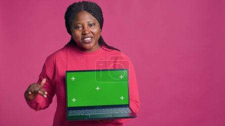Junge Dame mit afrikanisch-amerikanischer Ethnizität greift nach Laptop und präsentiert isolierte Chromakey-Vorlage. Minicomputer mit grünem Bildschirm-Attrappe, gehalten von einer schönen schwarzen Frau.