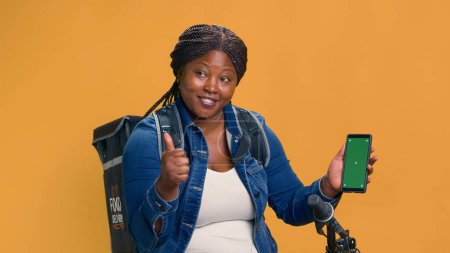 Téléchargez les photos : African American bike messenger tenant verticalement un appareil mobile avec écran vert isolé. Courrier féminin montrant l'application de livraison sur smartphone avec modèle chromakey. - en image libre de droit