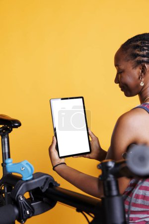 Photo for Detailed view of african american lady grasps device with blank copyspace display and having damaged bicycle nearby. Black woman holds tablet with chromakey mockup screen for bike maintenance. - Royalty Free Image