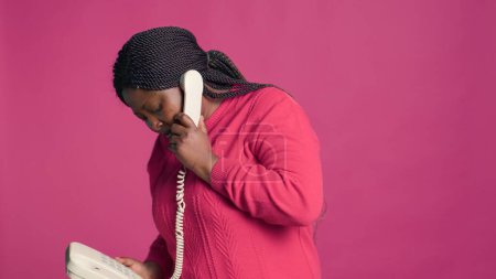 Foto de Retrato de mujer negra en suéter rosa de pie con teléfono móvil decepcionándose por la discusión. Joven afroamericana dama teniendo seria charla a través de teléfono fijo. - Imagen libre de derechos