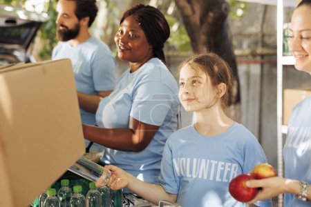 Photo for Photo focus on young volunteer girl sharing water and neccesities to poor needy and homeless people. Image showing charity workers giving fresh fruits and donation boxes to the less fortunate. - Royalty Free Image